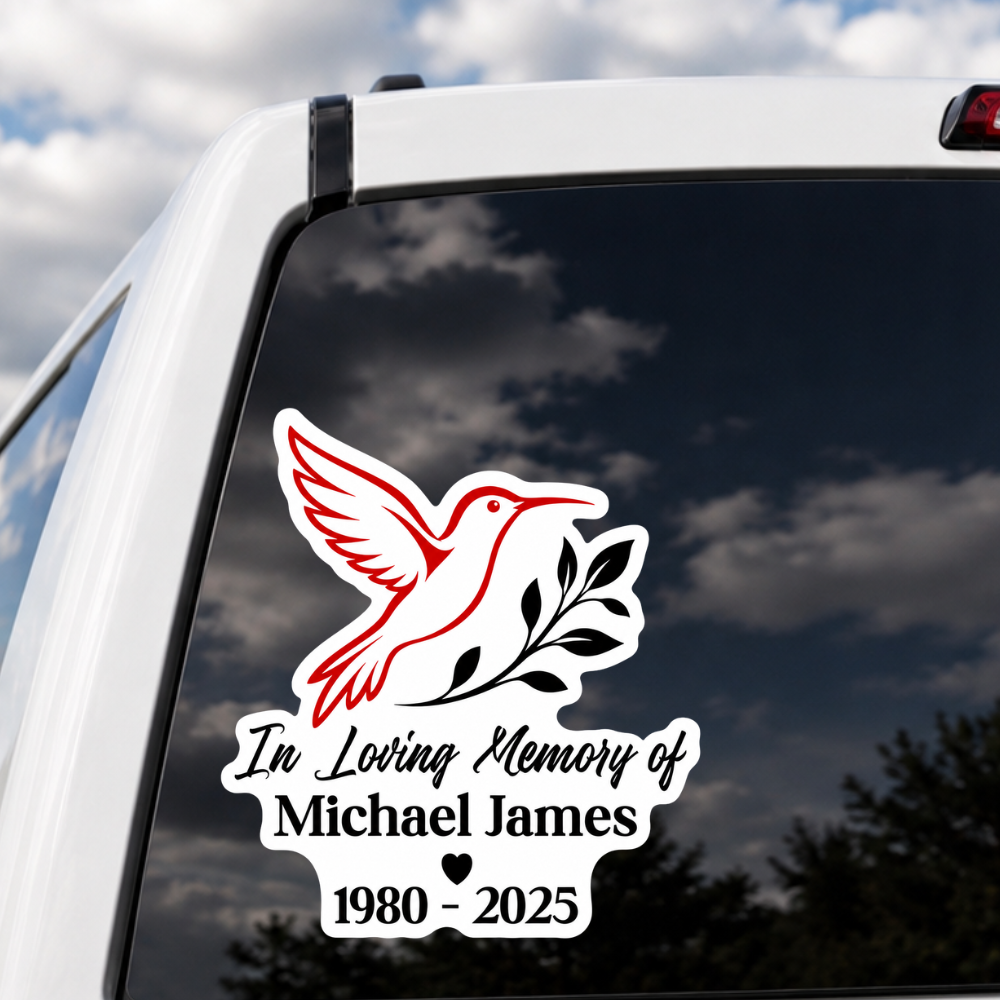 A memorial car window decal featuring a red and white hummingbird with black leaves and the text 'In Loving Memory applied to the back of a white vehicle.